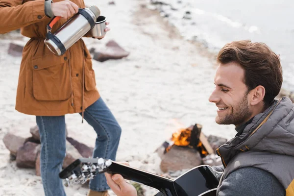 Jeune homme souriant jouant de la guitare acoustique près de la femme versant boisson chaude de thermos — Photo de stock