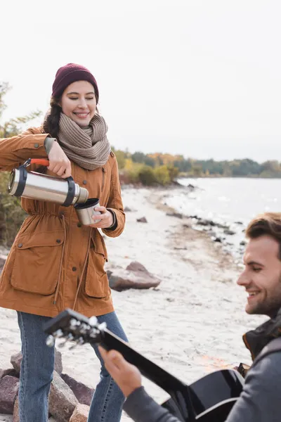 Cheerful woman pouring drink from thermos near blurred man playing guitar on sea coast — Stock Photo