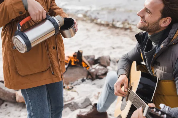 Glücklicher Mann spielt Gitarre neben Frau, die Getränk aus Thermoskanne einschenkt — Stockfoto