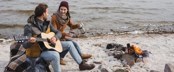 Happy woman holding thermos near man playing guitar while sitting on sea coast near bonfire, banner — Stock Photo