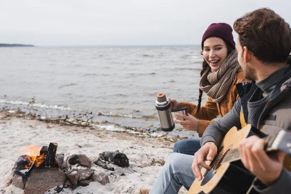 Hombre borroso con guitarra y mujer feliz con termos sentado en la hoguera en la costa del mar - foto de stock