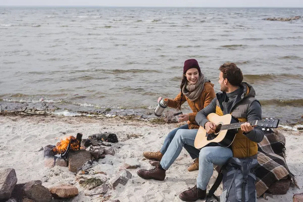 Hombre tocando la guitarra a la mujer feliz con termos mientras está sentado cerca de la hoguera en la costa del mar - foto de stock