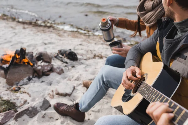 Частковий вид чоловіка, який грає на гітарі біля жінки з термосом на березі річки — стокове фото