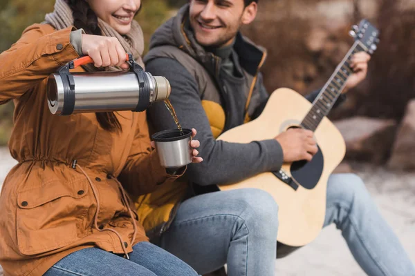 Vista cortada de mulher sorridente derramando chá de garrafa térmica perto de homem borrado tocando guitarra — Fotografia de Stock