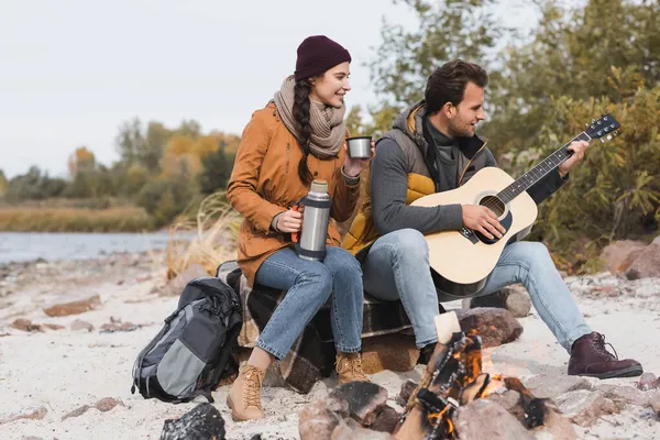 Mulher com termo e homem com guitarra acústica sentado perto da fogueira — Fotografia de Stock