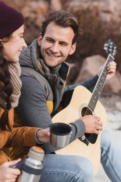 Homem feliz tocando guitarra acústica para mulher borrada com termo — Fotografia de Stock