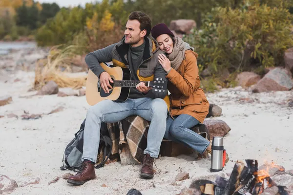 Giovane uomo che suona la chitarra vicino alla donna felice vicino al falò durante la sosta a piedi — Foto stock