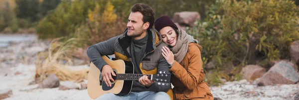 Mujer alegre con los ojos cerrados abrazando al hombre tocando la guitarra al aire libre, bandera - foto de stock