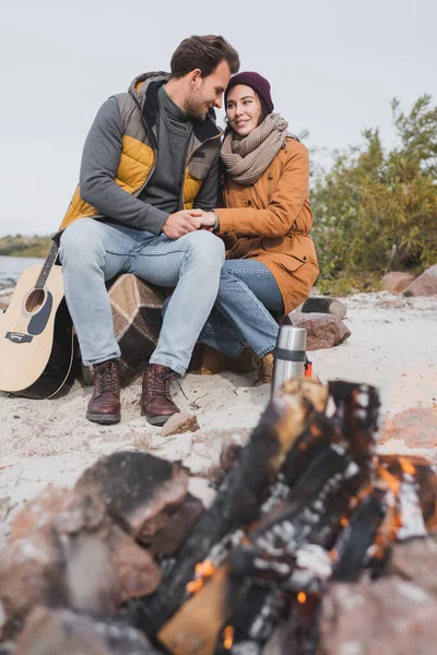 Glückliches Paar im Herbst-Outfit Händchen haltend am verschwommenen Lagerfeuer sitzend — Stockfoto