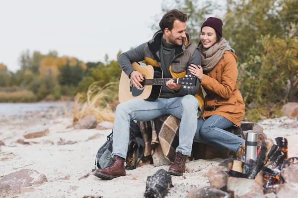 Hombre feliz tocando la guitarra cerca de la mujer mientras está sentado en piedras y manta durante el paseo de otoño - foto de stock