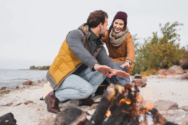 Young hikers in autumn clothes warming near blurred bonfire on riverside — Stock Photo