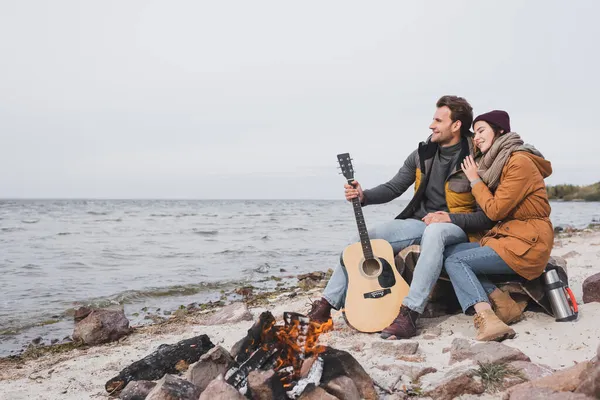 Happy couple with guitar and thermos sitting near bonfire on sea coast during halt in walk — Stock Photo