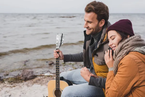 Mujer feliz con los ojos cerrados y joven con la guitarra sentado cerca del río - foto de stock