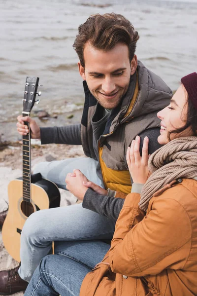 Cheerful woman touching shoulder of smiling man with guitar while sitting near lake — Stock Photo