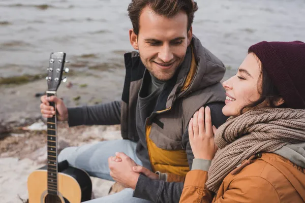 Joyful couple in autumn clothes looking at each other while sitting on sea shore — Stock Photo