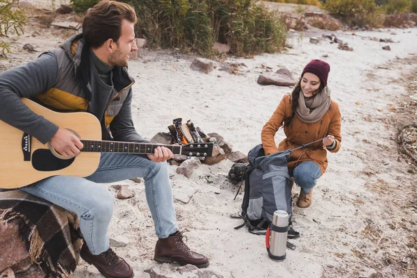 Uomo che suona la chitarra alla donna sorridente vicino zaino e thermos — Foto stock