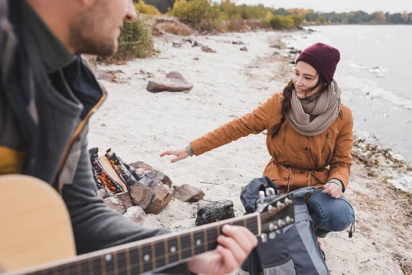 Uomo sfocato suonare la chitarra vicino sorridente donna riscaldamento vicino al falò — Foto stock