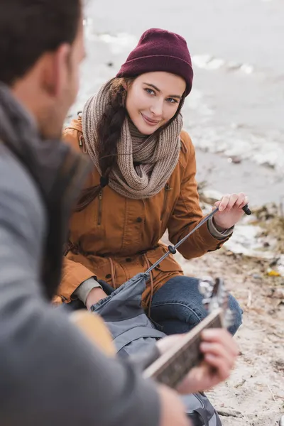 Mulher feliz abrindo mochila enquanto olha para o homem desfocado tocando guitarra — Fotografia de Stock