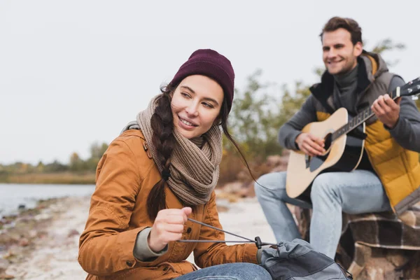 Mujer alegre mirando hacia otro lado y sosteniendo la mochila cerca del hombre borroso tocando la guitarra acústica - foto de stock