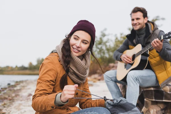 Fröhliche Frau schaut weg und hält Rucksack neben verschwommenem Mann, der Akustikgitarre spielt — Stockfoto