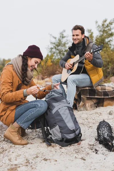 Smiling woman opening backpack near blurred man playing guitar during halt — Stock Photo