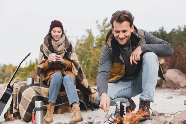 Femme heureuse assis avec tasse thermo sous couverture chaude près du petit ami et feu de joie — Photo de stock