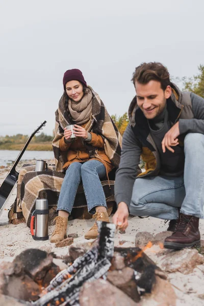 Young man near bonfire, and woman sitting with thermo cup under plaid blanket — Stock Photo