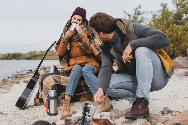 Mujer joven bebiendo de la taza termo cerca de novio y hoguera en la costa del río - foto de stock