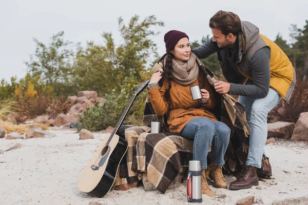 Junge Frau sitzt mit Thermoskanne in der Nähe von Thermoskanne und Gitarre, während ihr Freund sie mit einer warmen Decke bedeckt — Stockfoto