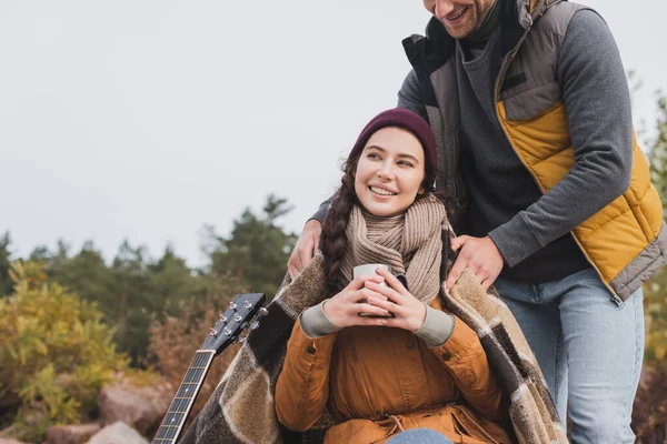 Cheerful woman holding thermo cup while boyfriend covering her with warm blanket outdoors — Stock Photo