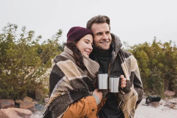 Pareja alegre, envuelta en manta caliente a cuadros, sosteniendo tazas termo al aire libre - foto de stock