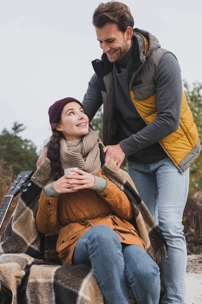 Young man in autumn clothes covering girlfriend with warm blanket during halt in walk — Stock Photo