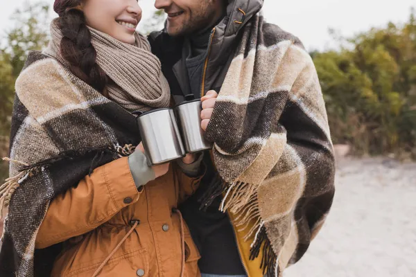 Partial view of young hikers with thermo cups wrapping in checkered blanket outdoors — Stock Photo