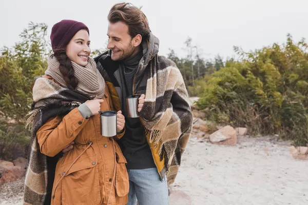 Feliz pareja joven sosteniendo vasos termo bajo una manta caliente durante el paseo de otoño - foto de stock