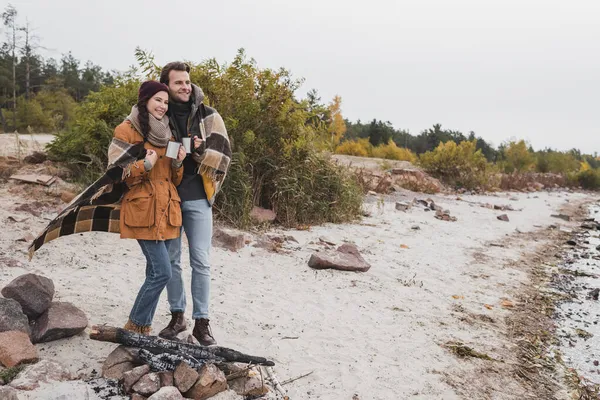 Casal feliz com copos térmicos de pé perto da fogueira sob manta xadrez — Fotografia de Stock