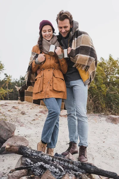 Turistas sonrientes en traje de otoño de pie con copas termo cerca de la hoguera - foto de stock