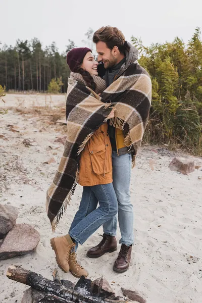 Alegre pareja de excursionistas abrazando mientras se calienta bajo manta a cuadros al aire libre - foto de stock