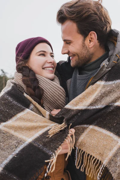 Casal alegre, coberto com manta quente xadrez, sorrindo um para o outro ao ar livre — Fotografia de Stock