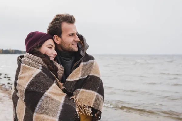 Giovane e felice coppia coperta di coperta plaid guardando lontano vicino al mare — Foto stock