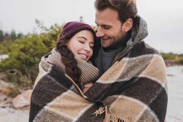 Feliz joven pareja calentamiento en a cuadros manta al aire libre - foto de stock