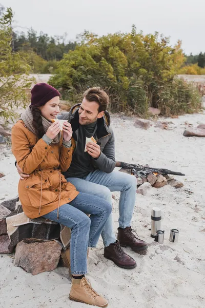 Happy couple of hikers eating sandwiches while sitting on stones and blanket outdoors — Stock Photo