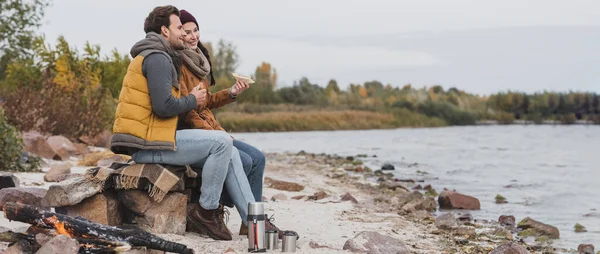 Young couple with sandwiches sitting on stones and blanket near thermo cups on river shore, banner — Stock Photo