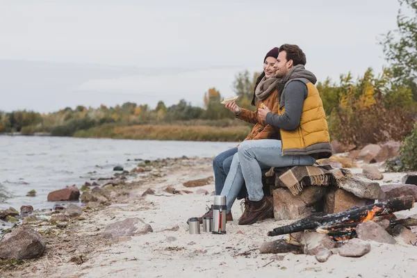 Feliz pareja en ropa de otoño mirando el río mientras se sienta con sándwiches - foto de stock