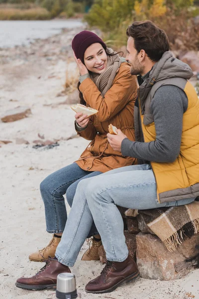 Sonriente pareja sentada en manta a cuadros con sándwiches durante la parada en otoño a pie - foto de stock