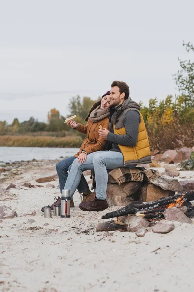 Couple heureux avec des sandwichs assis sur des pierres et une couverture près du thermos et du feu de joie — Photo de stock