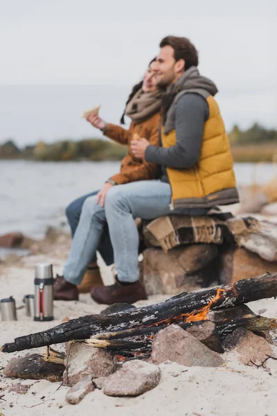 Blurred couple with sandwiches sitting near river and bonfire on coast — Stock Photo