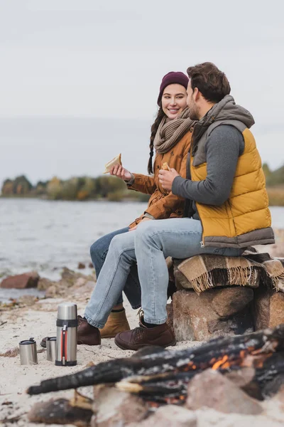 Счастливая пара с бутербродами, сидящими на камнях и теплым одеялом во время остановки на берегу реки — стоковое фото
