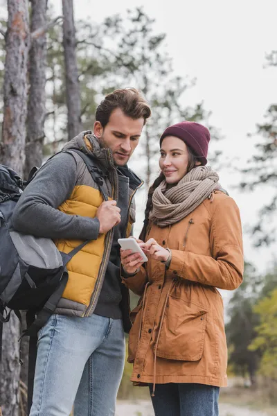 Mujer sonriente con smartphone y hombre con mochila caminando en el bosque - foto de stock