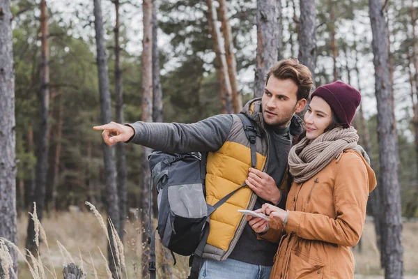 Hombre señalando con el dedo cerca de la mujer con el teléfono móvil mientras camina en el bosque - foto de stock