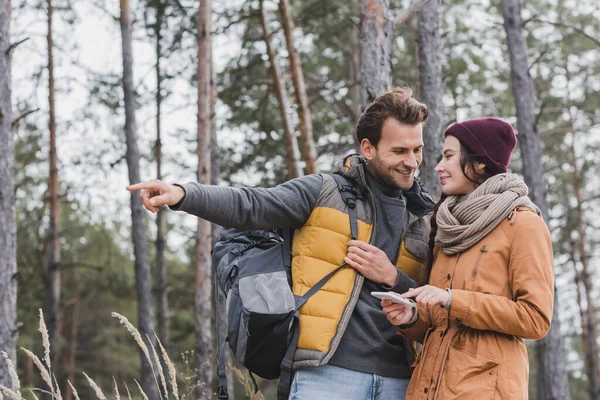 Hombre sonriente con mochila apuntando con el dedo cerca de la novia utilizando el teléfono inteligente en el bosque - foto de stock